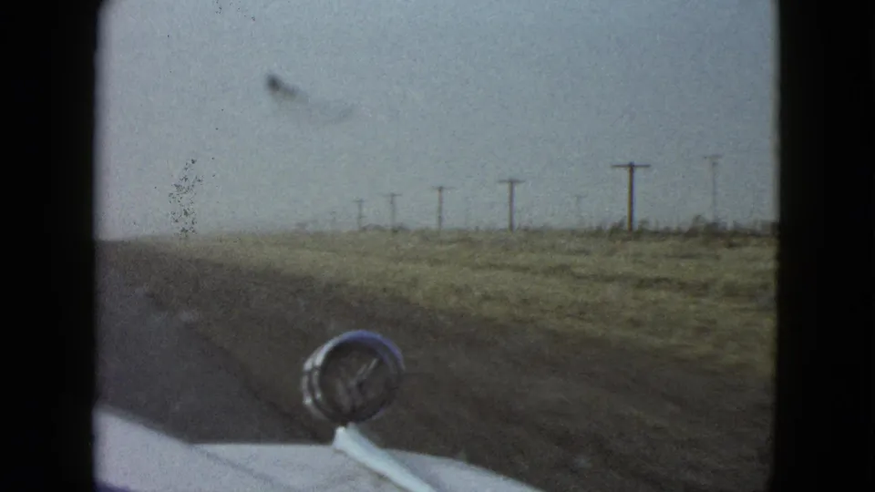 1958 Las Vegas-area home-movie frame looking from a moving car past a chrome hood ornament toward a flat roadside with utility poles.