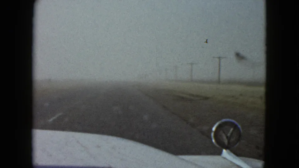 Passenger-view frame from a 1958 Las Vegas-area drive, showing the road ahead, car hood ornament, roadside poles, and a small bird in the sky.