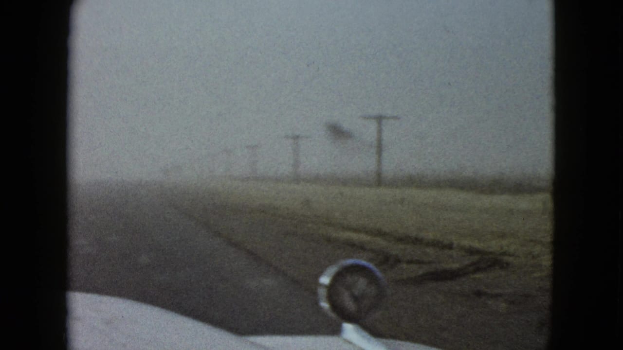 1958 home-movie view near Las Vegas shows a foggy rural road, roadside utility poles, and a car hood ornament through the windshield.