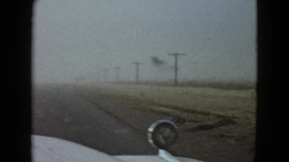 1958 home-movie view near Las Vegas shows a foggy rural road, roadside utility poles, and a car hood ornament through the windshield.