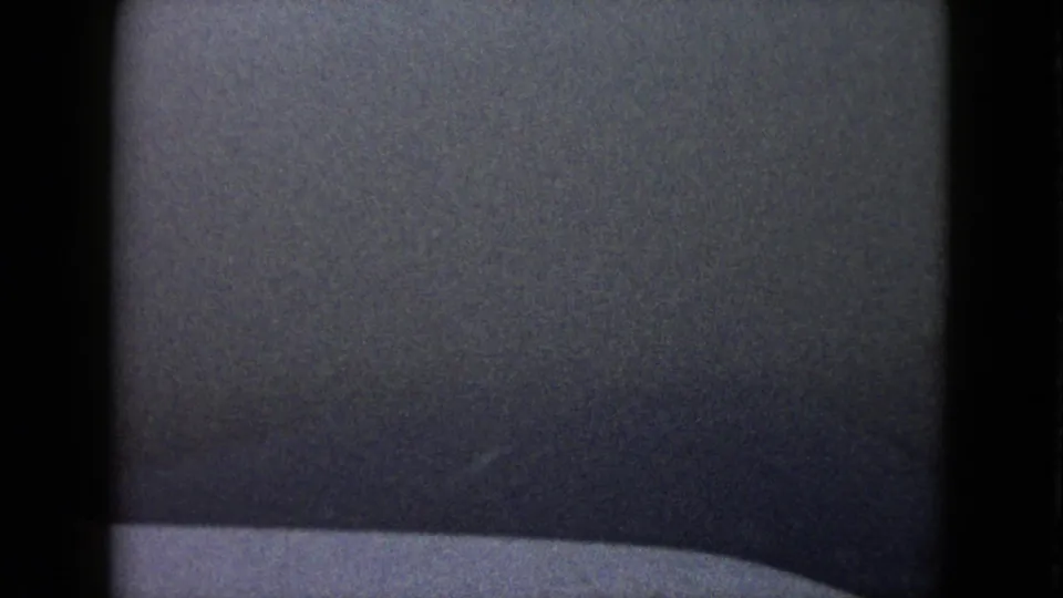 1958 8mm frame with a dark, indistinct field, pale lower edge, grain, and gate borders suggesting a transition or obscured vehicle view.