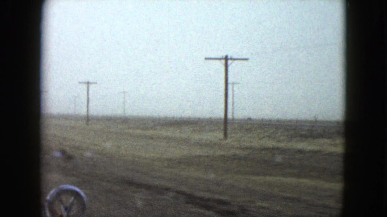 1958 Nevada car-window view of flat roadside terrain, utility poles, overcast sky, and the edge of the vehicle hood.