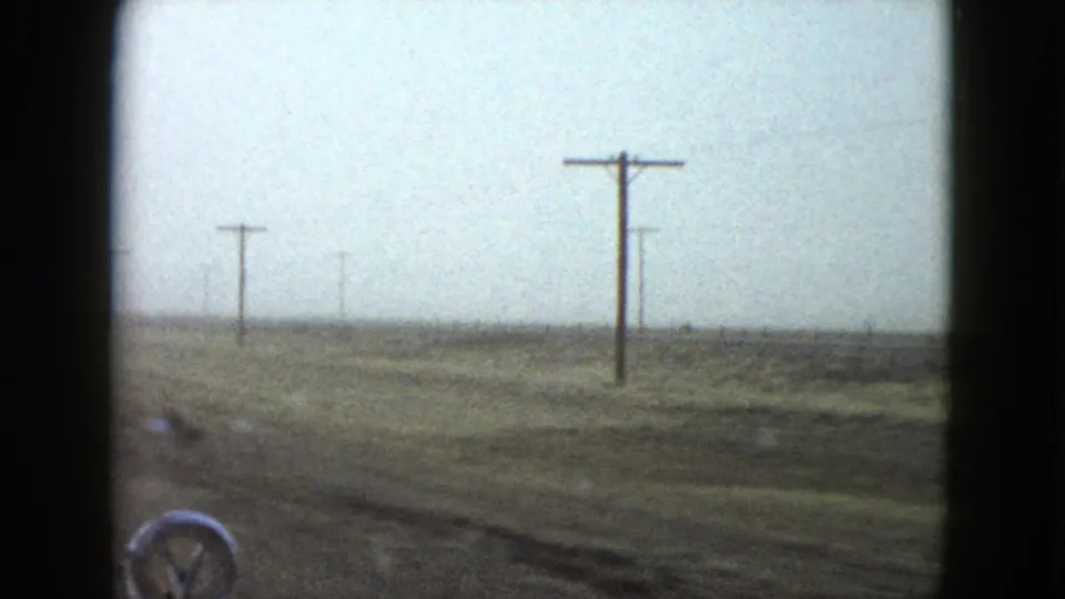 1958 Nevada car-window view of flat roadside terrain, utility poles, overcast sky, and the edge of the vehicle hood.