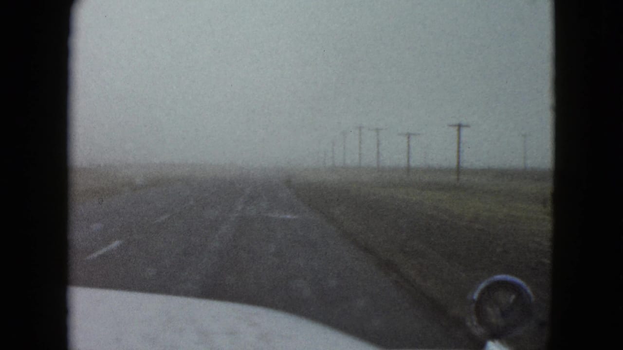 Forward-looking 1958 Nevada highway view from inside a car, with road markings, utility poles, and muted overcast light.