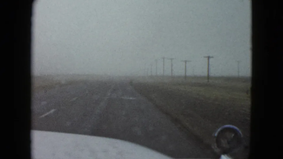 Forward-looking 1958 Nevada highway view from inside a car, with road markings, utility poles, and muted overcast light.