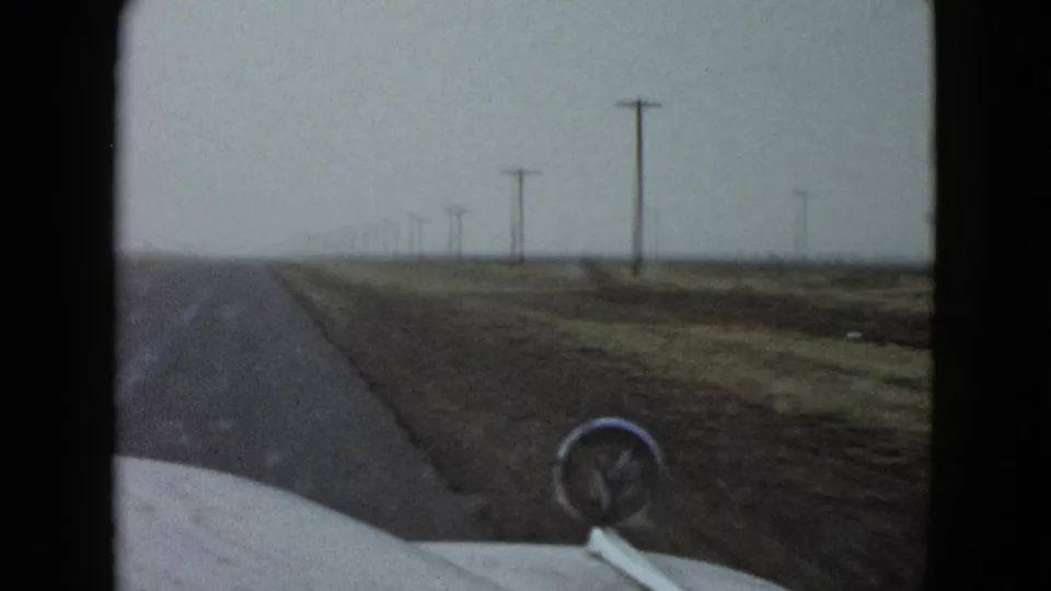 1958 home-movie view from a car on a straight road near Las Vegas, with flat fields and utility poles receding into the distance.