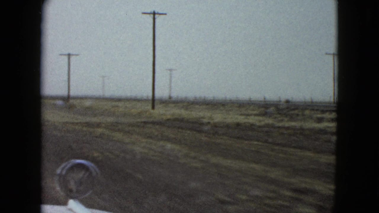 Roadside view from a moving car with utility poles, open fields, and a car hood ornament in front.
