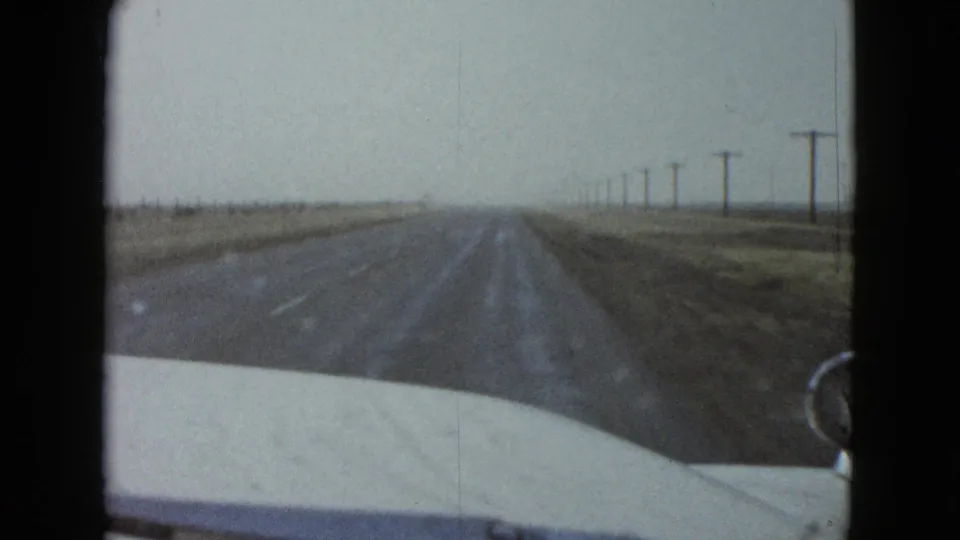 1958 8mm travel footage shows the road ahead from inside a moving car, with open land and utility poles fading into haze.