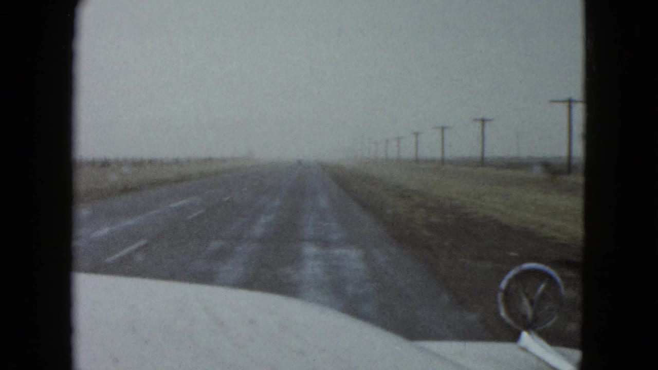 1958 Las Vegas-area home-movie view from inside a moving car on a straight rural road with utility poles receding ahead.