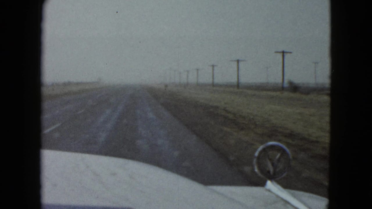 Forward windshield view on a rural Las Vegas road in 1958, showing the car hood, hood ornament, open land, and utility poles.