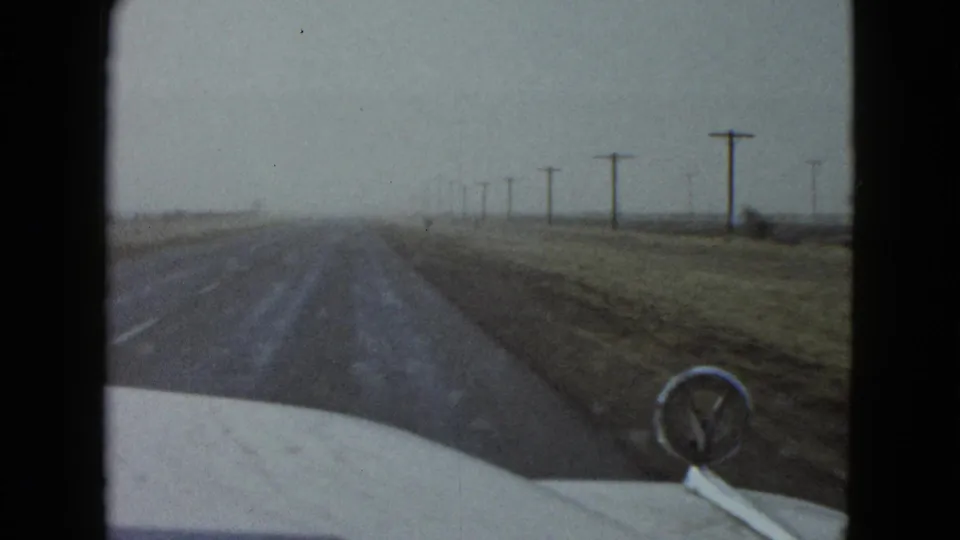 Forward windshield view on a rural Las Vegas road in 1958, showing the car hood, hood ornament, open land, and utility poles.