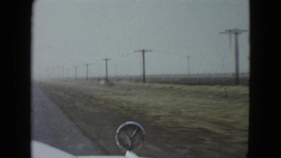 Side-forward car view from 1958 Las Vegas footage, with roadside grass, utility poles, open fields, and the hood ornament in frame.