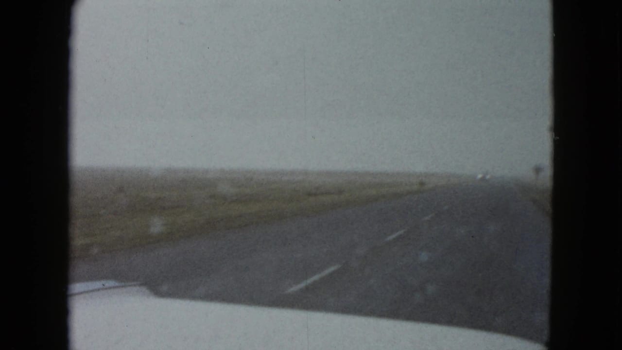 1958 8mm car POV near Las Vegas looking down an open highway bordered by low vegetation and distant utility poles.