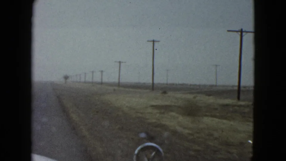1958 Las Vegas-area home-movie frame from inside a car, showing a straight roadside landscape with utility poles and a pale hood edge.