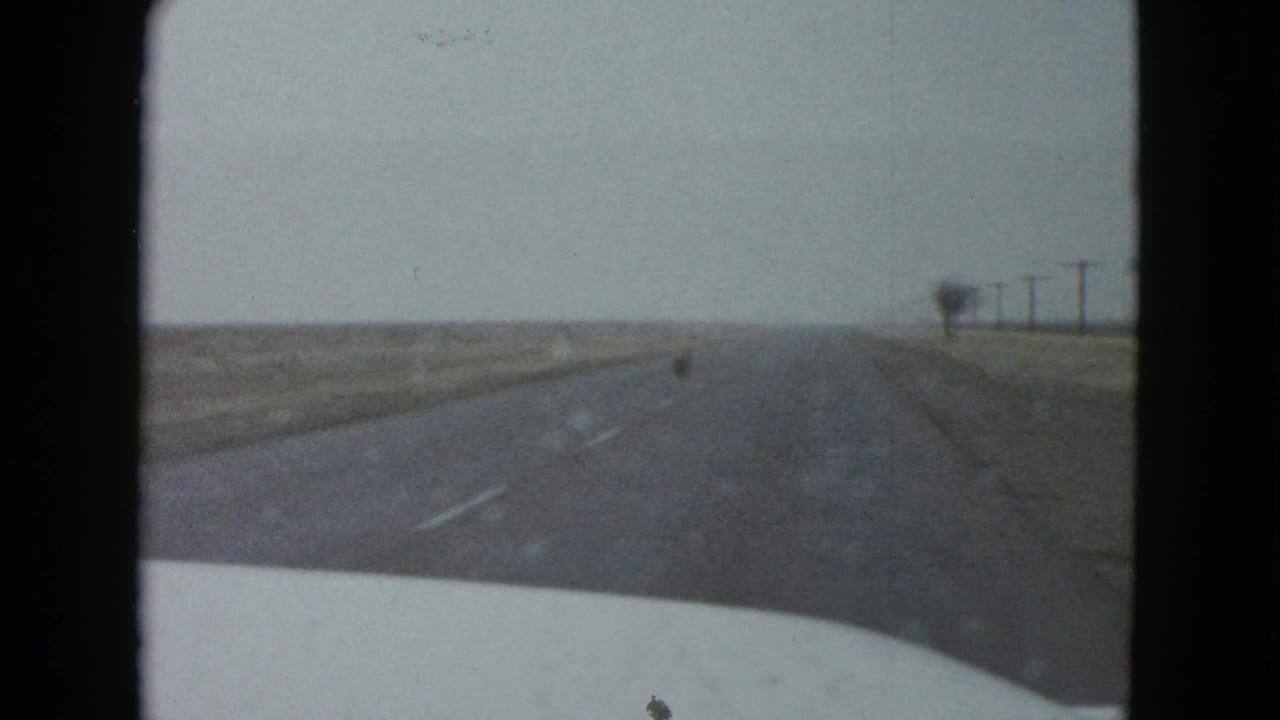Windshield view from a 1958 home movie near Las Vegas, with the road ahead, white hood, roadside fields, and distant utility poles.