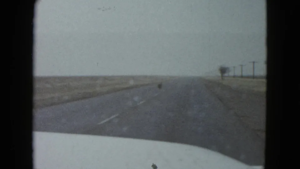 Windshield view from a 1958 home movie near Las Vegas, with the road ahead, white hood, roadside fields, and distant utility poles.