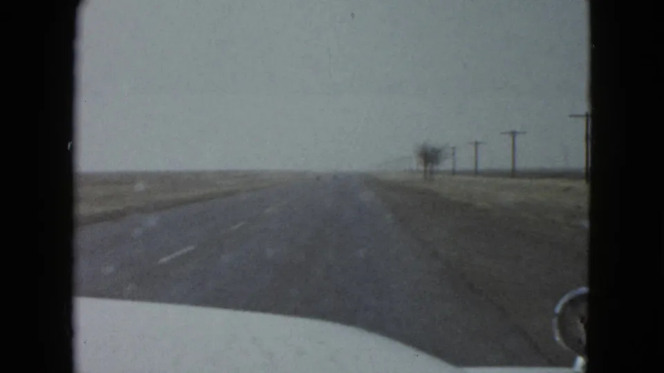1958 8mm windshield shot near Las Vegas, showing the car hood, straight road, open fields, and utility poles under a muted sky.