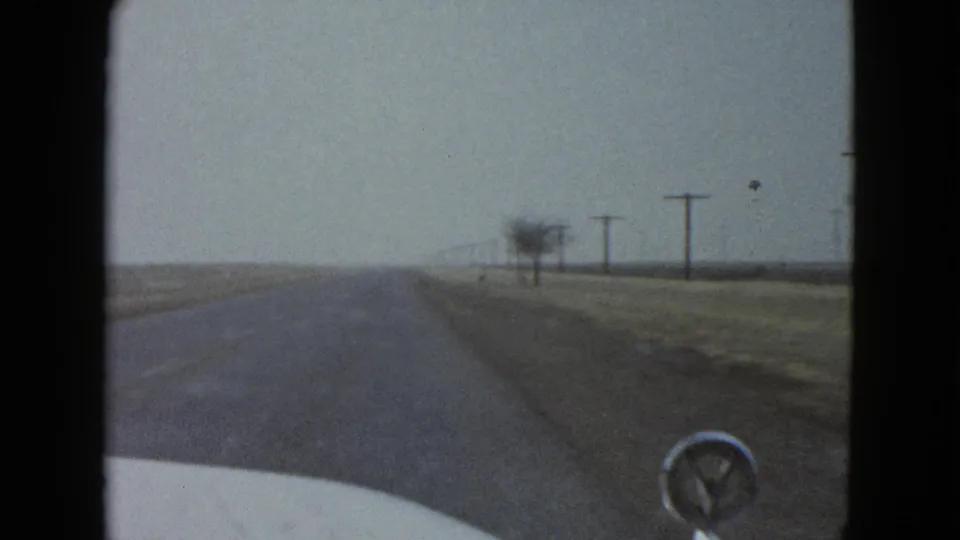 A bird passes in front of the windshield during a 1958 Las Vegas-area driving shot, with the road and utility poles visible beyond.