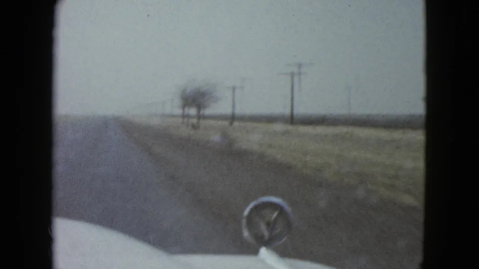 1958 Las Vegas-area home-movie view through a car windshield toward a rural road, roadside utility poles, dry grass, and a chrome hood ornament.