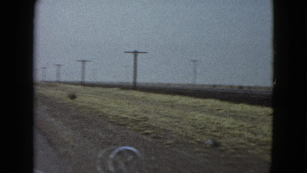 Windshield view from a 1958 home movie showing open rural Las Vegas-area roadside, utility poles, flat fields, and part of the car hood ornament.