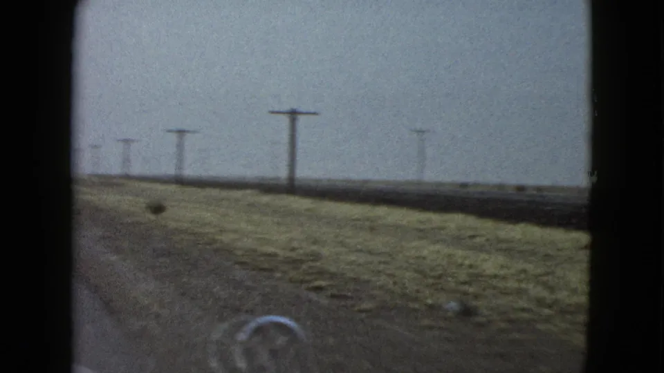 Windshield view from a 1958 home movie showing open rural Las Vegas-area roadside, utility poles, flat fields, and part of the car hood ornament.