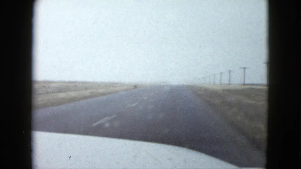 A bright 1958 Las Vegas-area driving view shows the road ahead through windshield glare, with dry roadside fields and utility poles fading into haze.