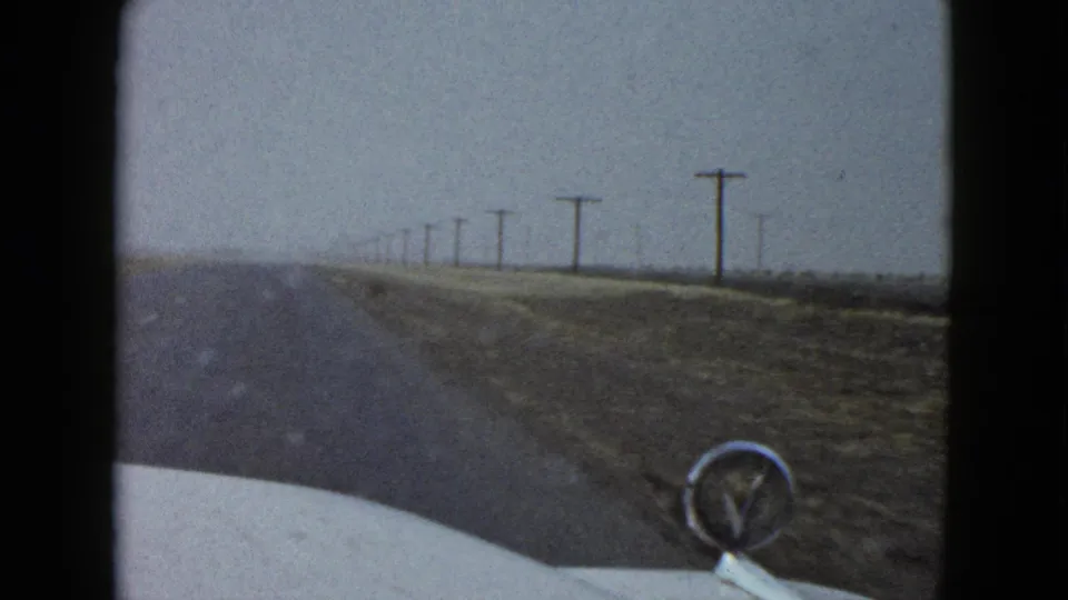 A 1958 home-movie frame from the car shows the hood ornament in front of a rural Las Vegas-area road, dry ground, and utility poles under haze.