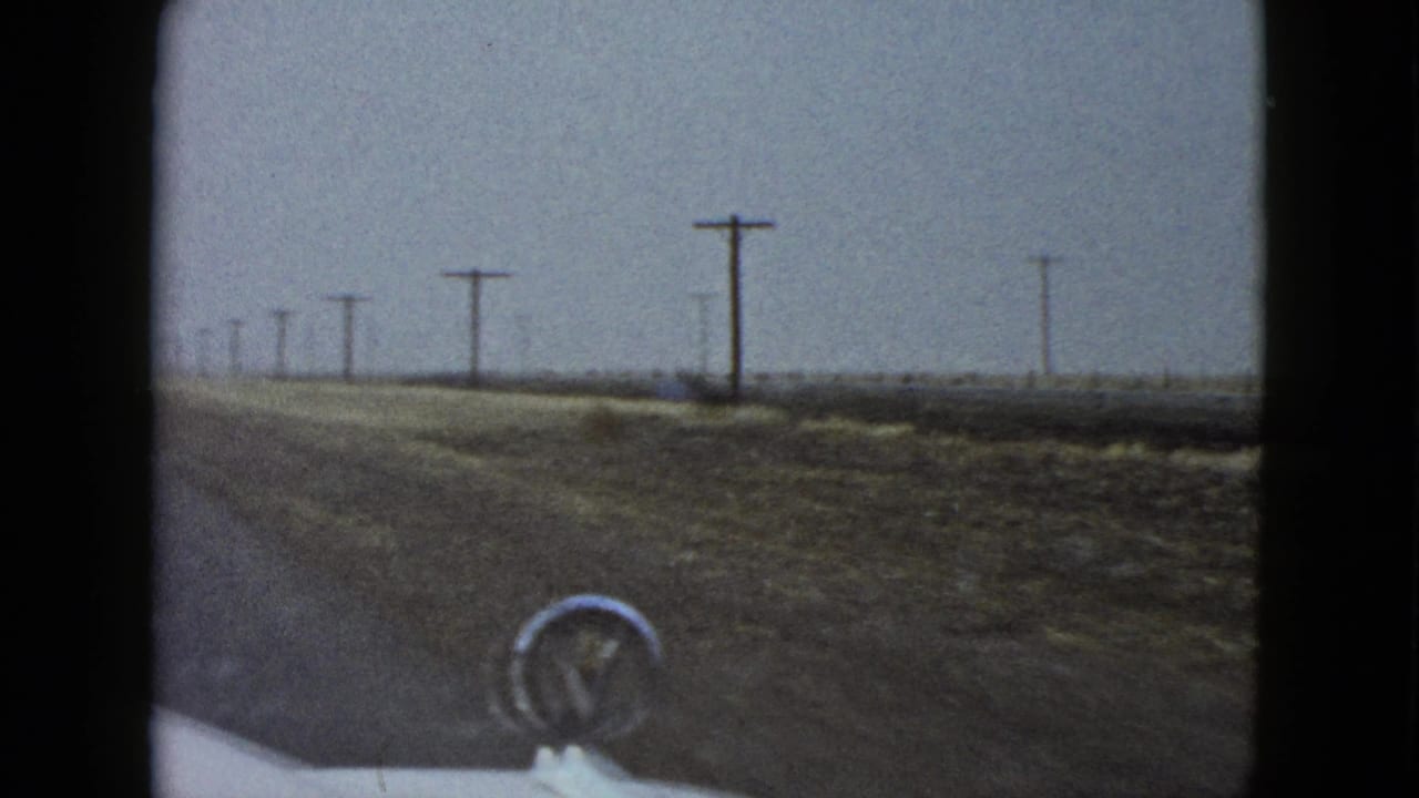 1958 8mm driver’s-view frame near Las Vegas showing a wet roadside, utility poles and a white car hood with a chrome ornament.