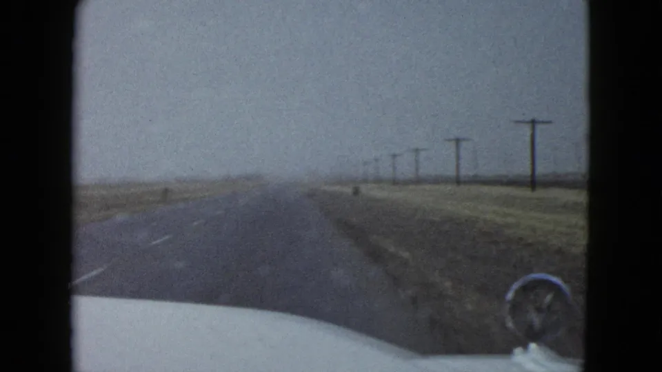 1958 home-movie frame from a moving car showing a wet highway, broad shoulder, utility poles and the car hood in foreground.
