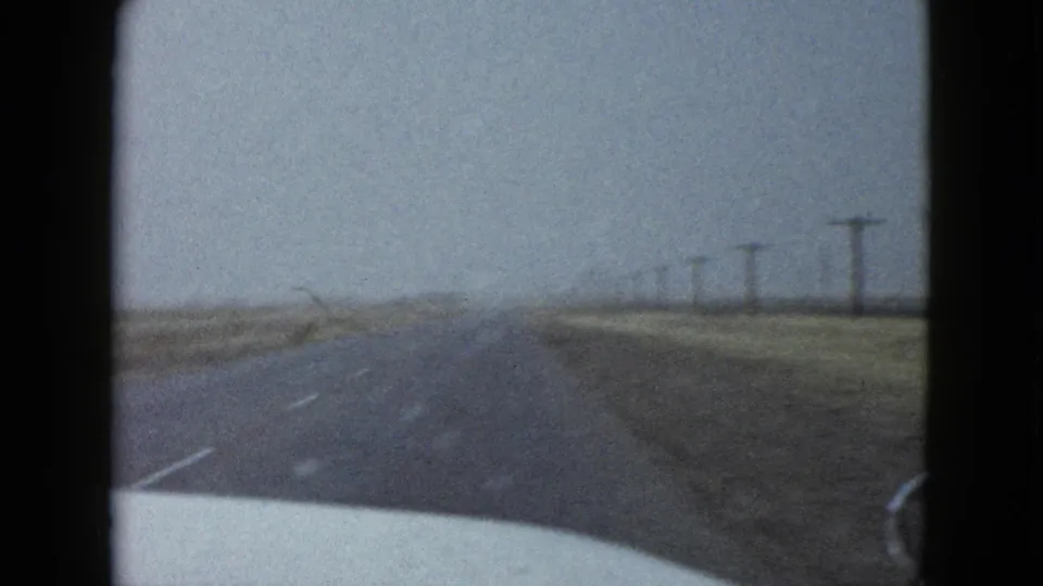 Driver’s-view 8mm frame near Las Vegas showing a damp highway fading into haze, bordered by open fields and utility poles.