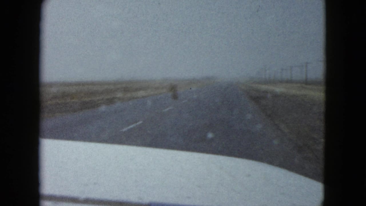 1958 Las Vegas-area 8mm frame looking through a car windshield toward a straight desert road, dry roadside land, distant utility poles, and a pale car hood at the bottom edge.