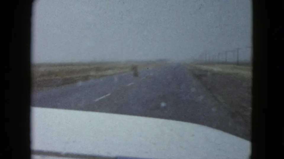1958 Las Vegas-area 8mm frame looking through a car windshield toward a straight desert road, dry roadside land, distant utility poles, and a pale car hood at the bottom edge.