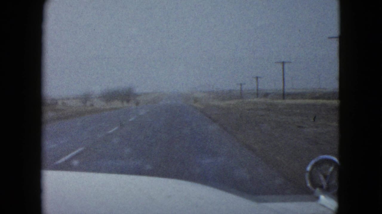 1958 Las Vegas-area road view from inside a moving car, with the car hood and side mirror visible as a tumbleweed or brush passes ahead on the desert highway.