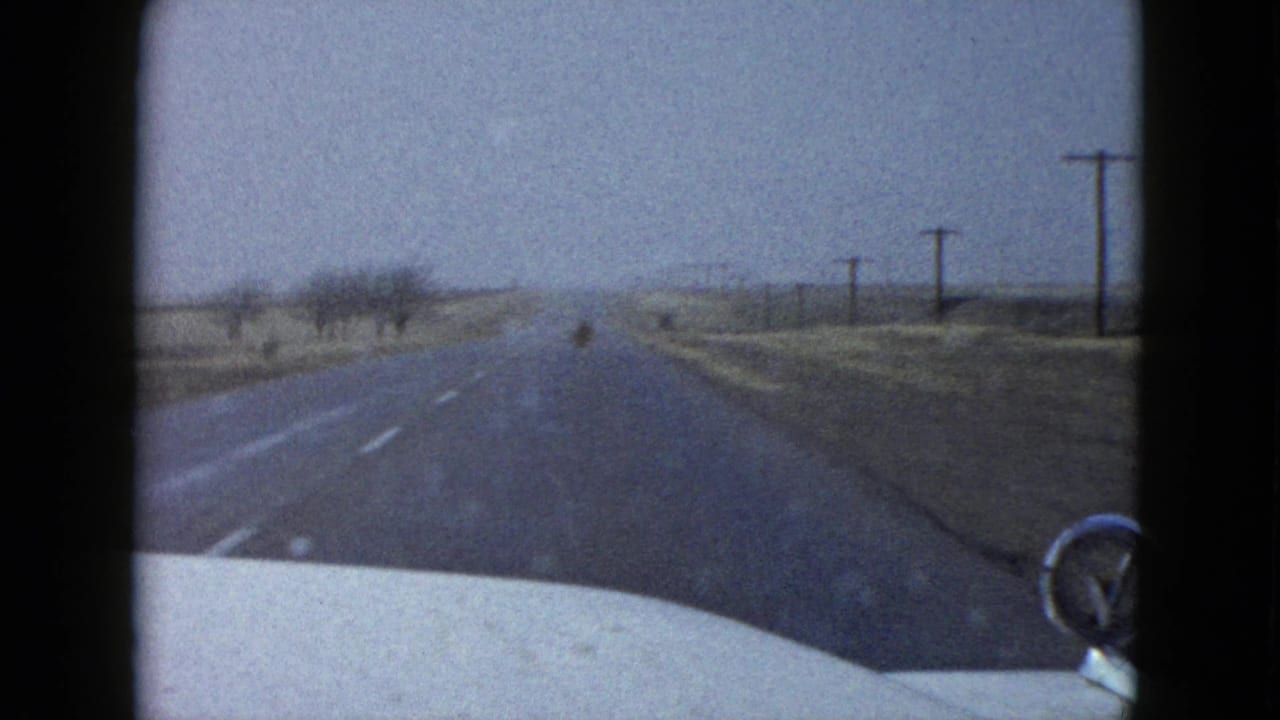 A 1958 Las Vegas-area 8mm frame shows a forward windshield view along a two-lane desert road, with the pale car hood, right-side mirror, utility poles, and dry roadside terrain visible.