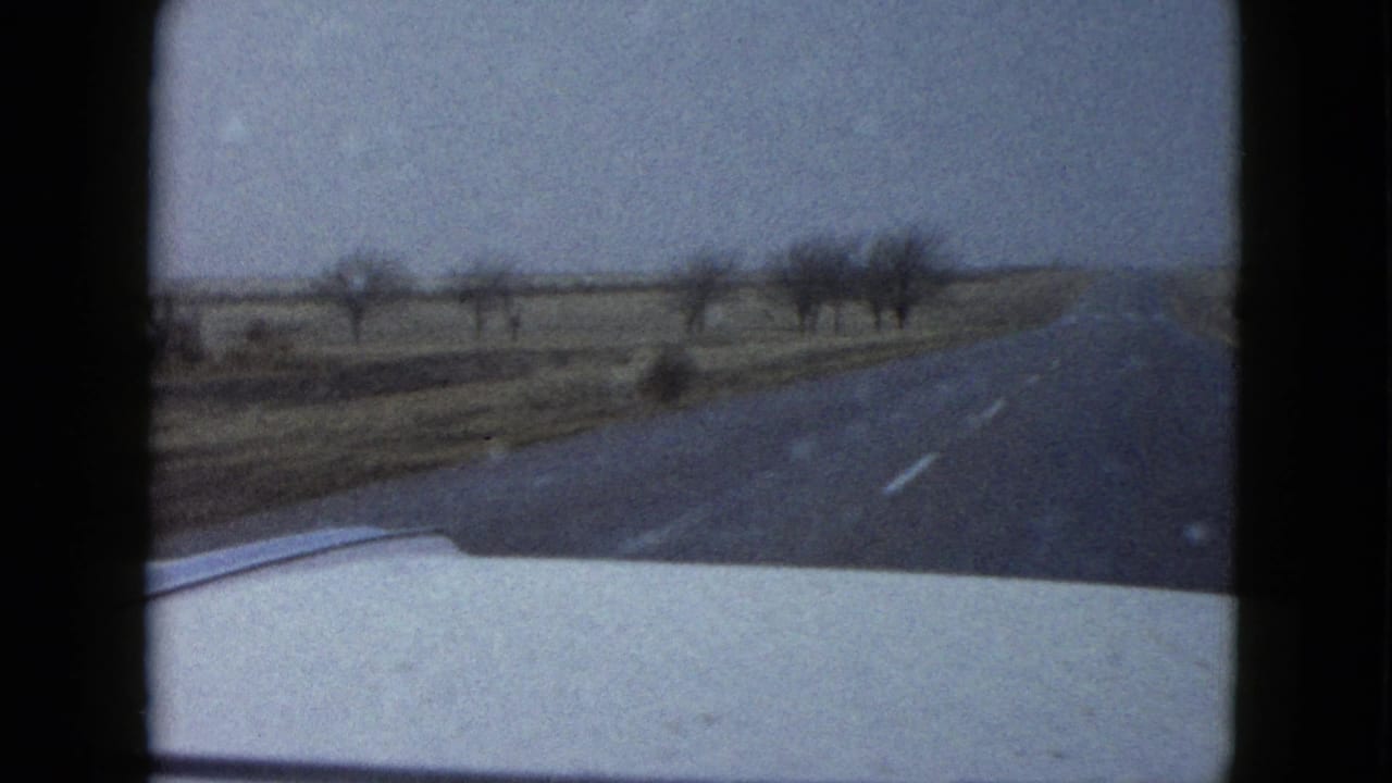 End frame from the 1958 Las Vegas-area clip, looking over the car hood as the desert road extends past scattered bare trees or brush and open roadside land.
