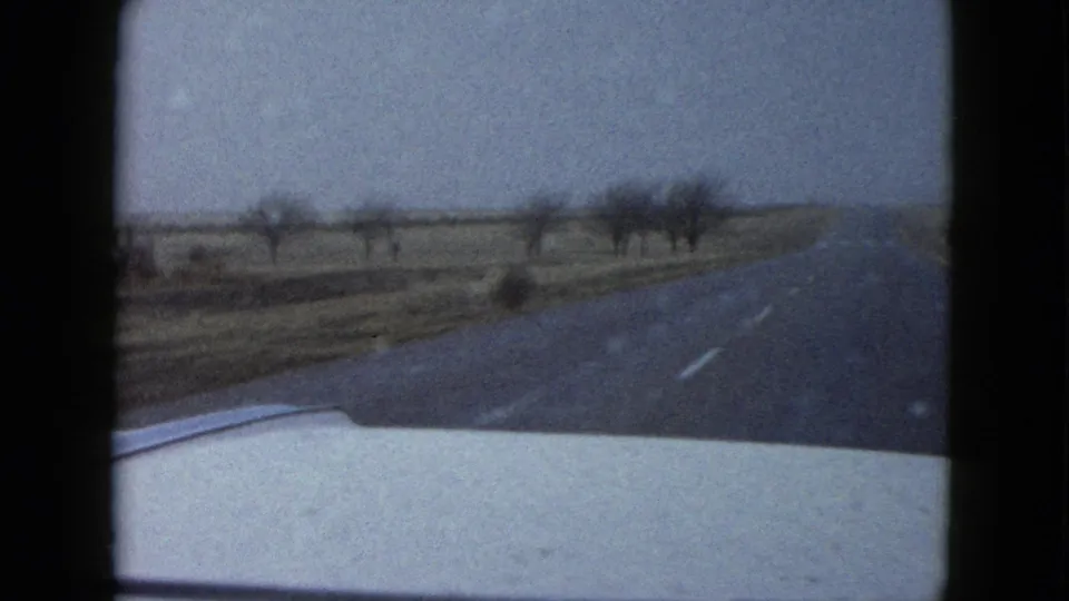 End frame from the 1958 Las Vegas-area clip, looking over the car hood as the desert road extends past scattered bare trees or brush and open roadside land.