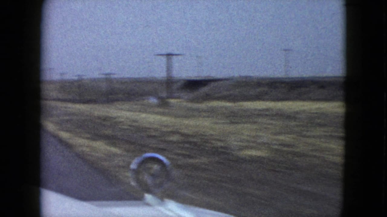 Roadside view from a moving car with a hood ornament in the foreground.