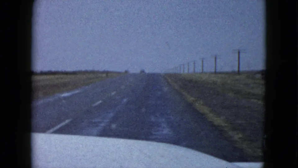 1950s rural highway view from a moving car, with wet or dark pavement, open roadside, and utility poles along the right side.