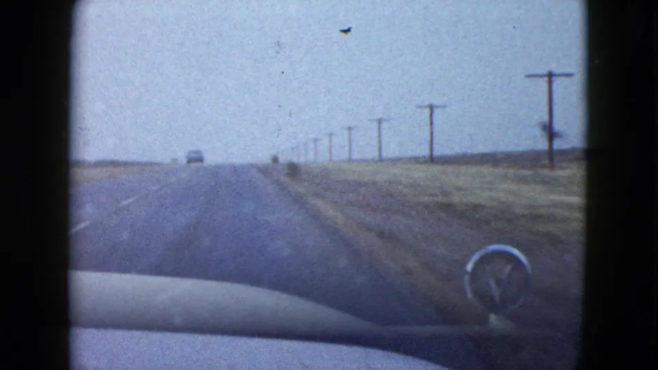 1958 Nevada highway footage showing the car hood and ornament, a distant vehicle, utility poles, and open roadside land.