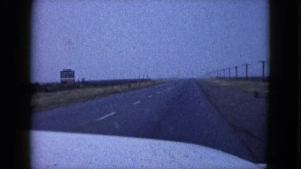 1958 Las Vegas-area road view from inside a moving car, showing a paved highway, open desert shoulder, utility poles, and a distant roadside sign.