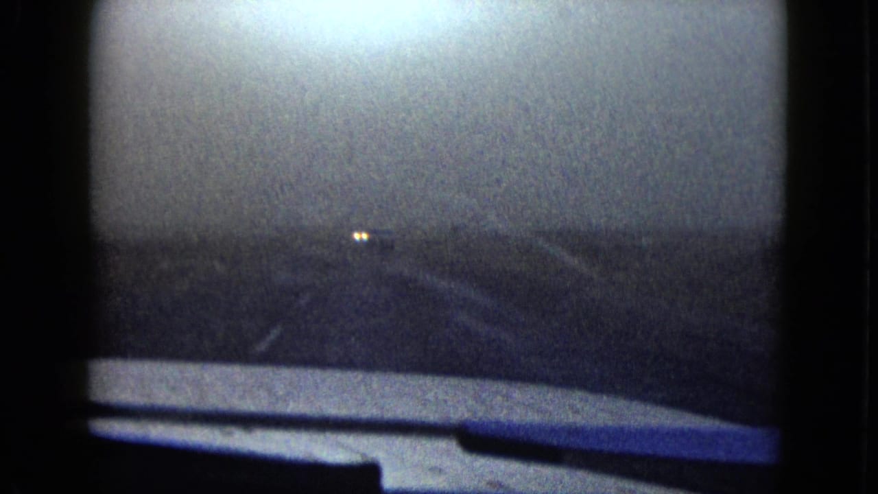 Dark view from inside a moving car with distant lights and a road edge.
