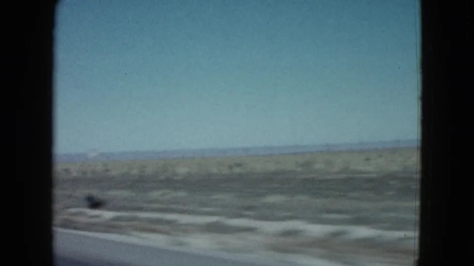 1958 Las Vegas-area desert road view filmed from inside a moving vehicle, with sandy terrain, sparse brush, and windshield edges visible.