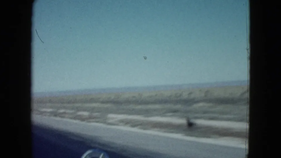 1958 Las Vegas-area highway view through a windshield, with dark vehicle hood edge, sandy roadside, low bushes, and distant horizon.