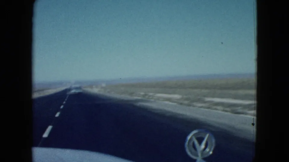1958 Las Vegas-area road footage showing an open asphalt lane, white markings, desert shoulder, and the rounded hood of the filming vehicle.