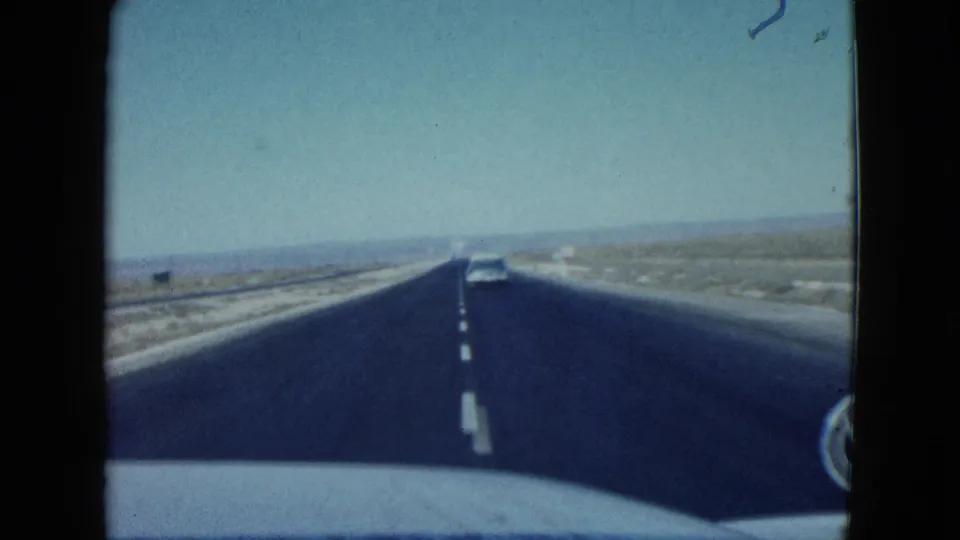 1958 Las Vegas-area highway scene from inside a moving car, with a distant vehicle ahead, lane markings, desert scrub, and pale sky.