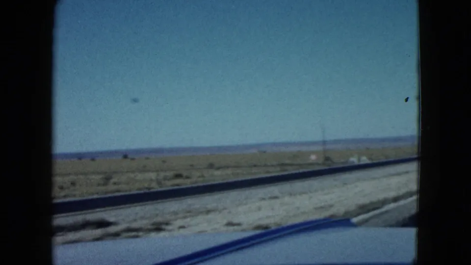 In-car 8mm view of a Nevada desert roadside, with low brush, distant hills, a utility pole, and blue car trim along the lower edge.