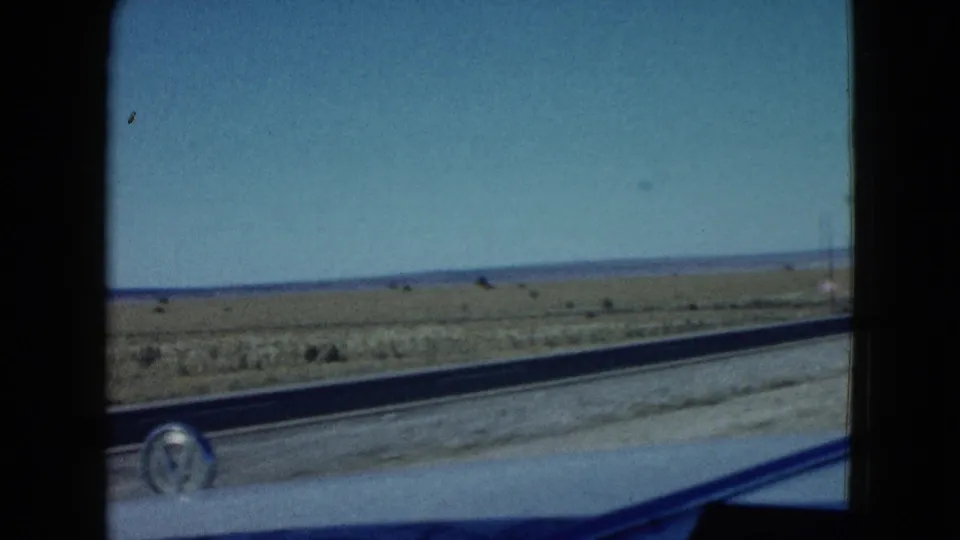 Late-frame in-car view near Las Vegas showing the highway edge, open desert, distant hills, utility pole, and car hood ornament.