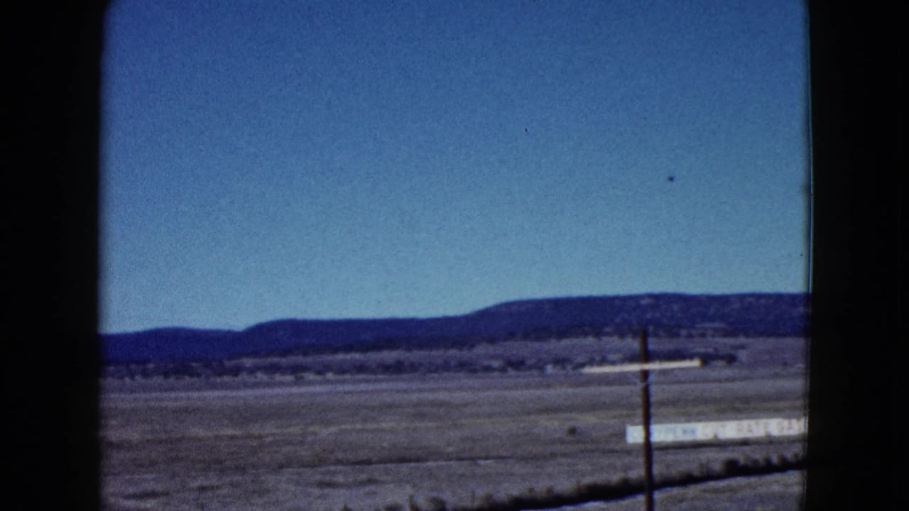 Flat desert field with distant mountains, clear blue sky, utility pole, and a pale roadside sign.