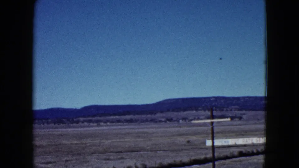 1958 Las Vegas-area 8mm frame showing open desert ground, a utility pole, a long pale roadside sign, and low mountains beneath clear blue sky.