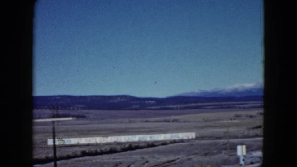 1958 Las Vegas-area 8mm frame showing roadside desert, a long pale sign, a pole, distant ridges, and snow-capped mountains on the horizon.